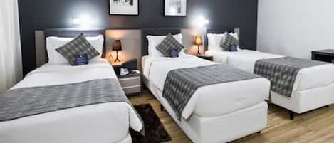 A hotel room with four single beds, a dark wall with two framed pictures, a small rug, and a wooden floor.