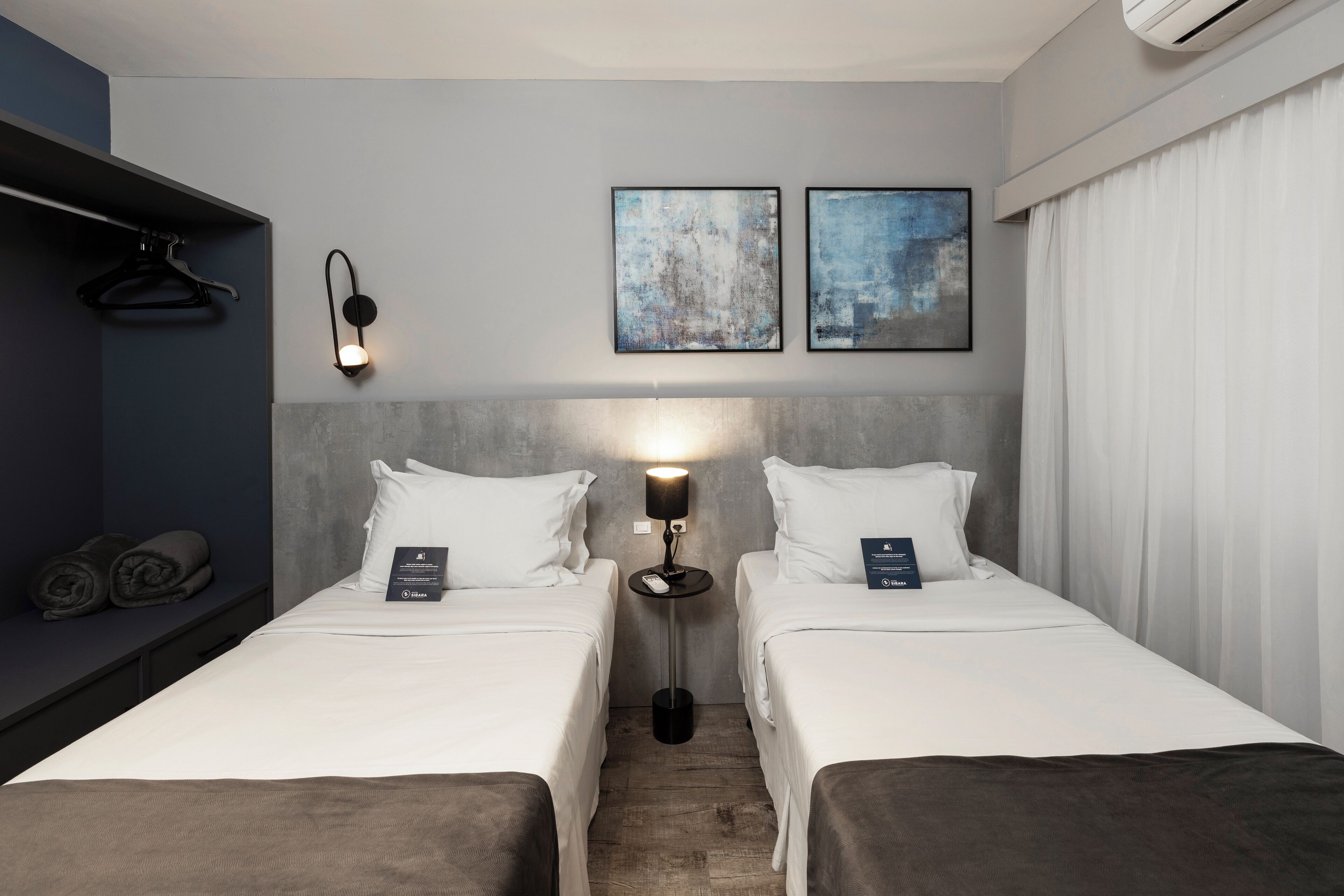 Habitación de hotel con dos camas, una mesita de noche con lámpara, una luz de pared y dos cuadros enmarcados.