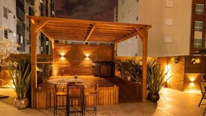 Una pérgola de madera al aire libre con una mesa redonda y sillas, rodeada de plantas en macetas.