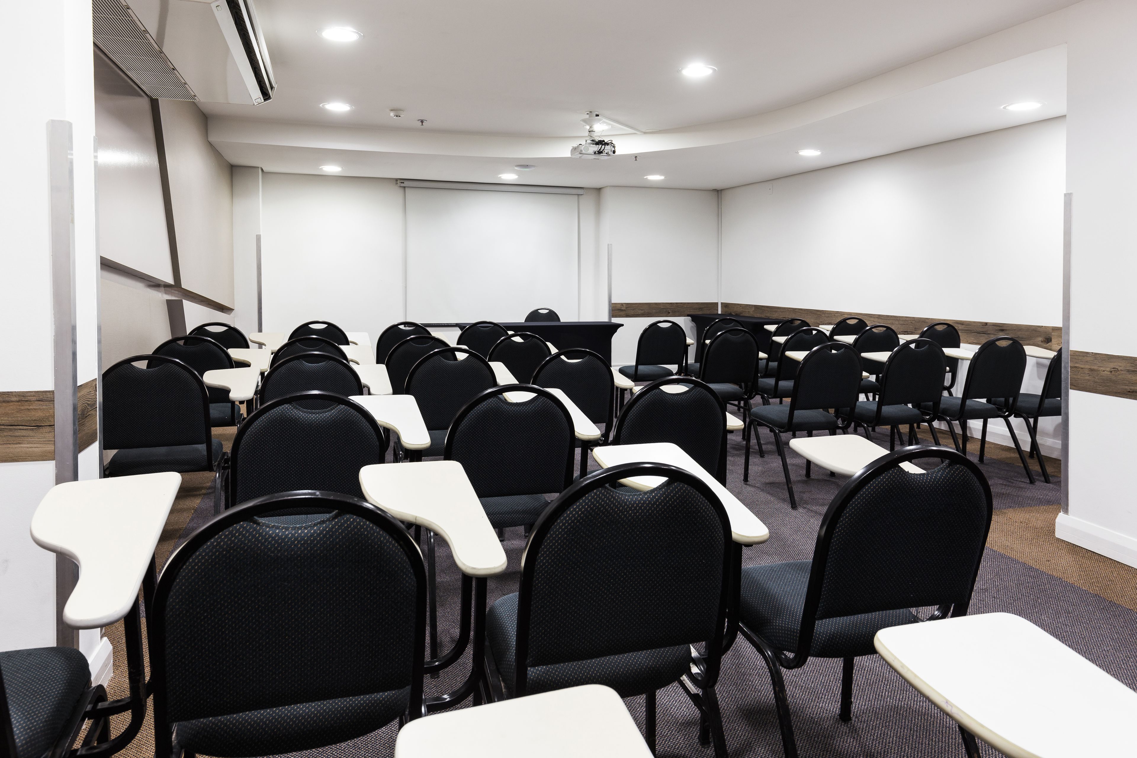 Sala de conferências com fileiras de cadeiras e mesas, paredes brancas e um projetor no teto.