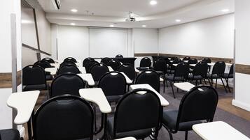 Sala de conferências com fileiras de cadeiras e mesas, paredes brancas e um projetor no teto.