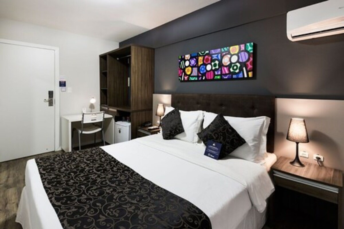 A hotel room with a bed, bedside tables, a desk, a wardrobe, and a colorful artwork on the wall.
