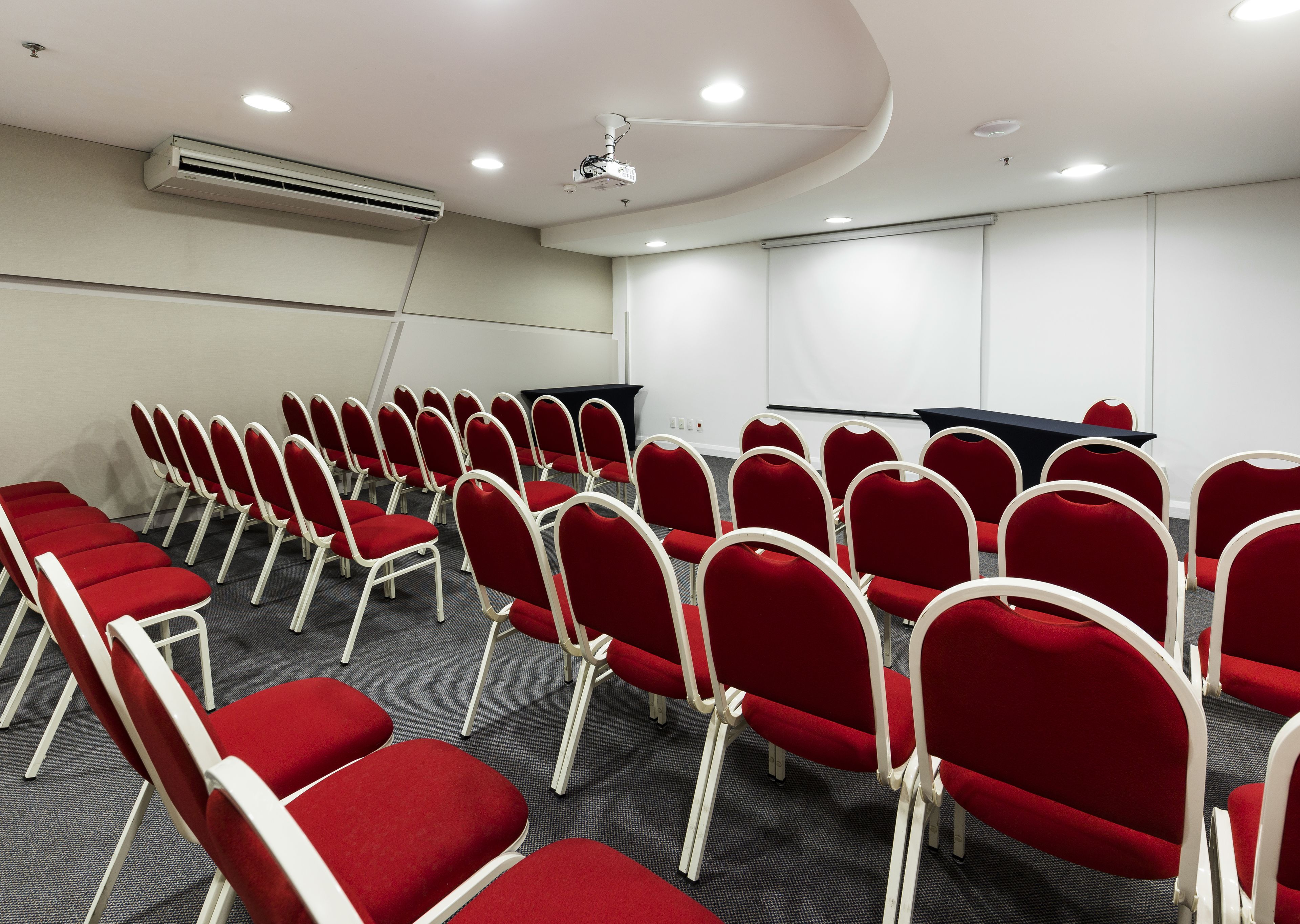 Une salle de conférence avec des rangées de chaises rouges, un écran de projection et une table noire.
