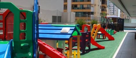 Un parque infantil con estructuras de juego coloridas, incluyendo toboganes y juegos de escalada, con edificios residenciales al fondo.