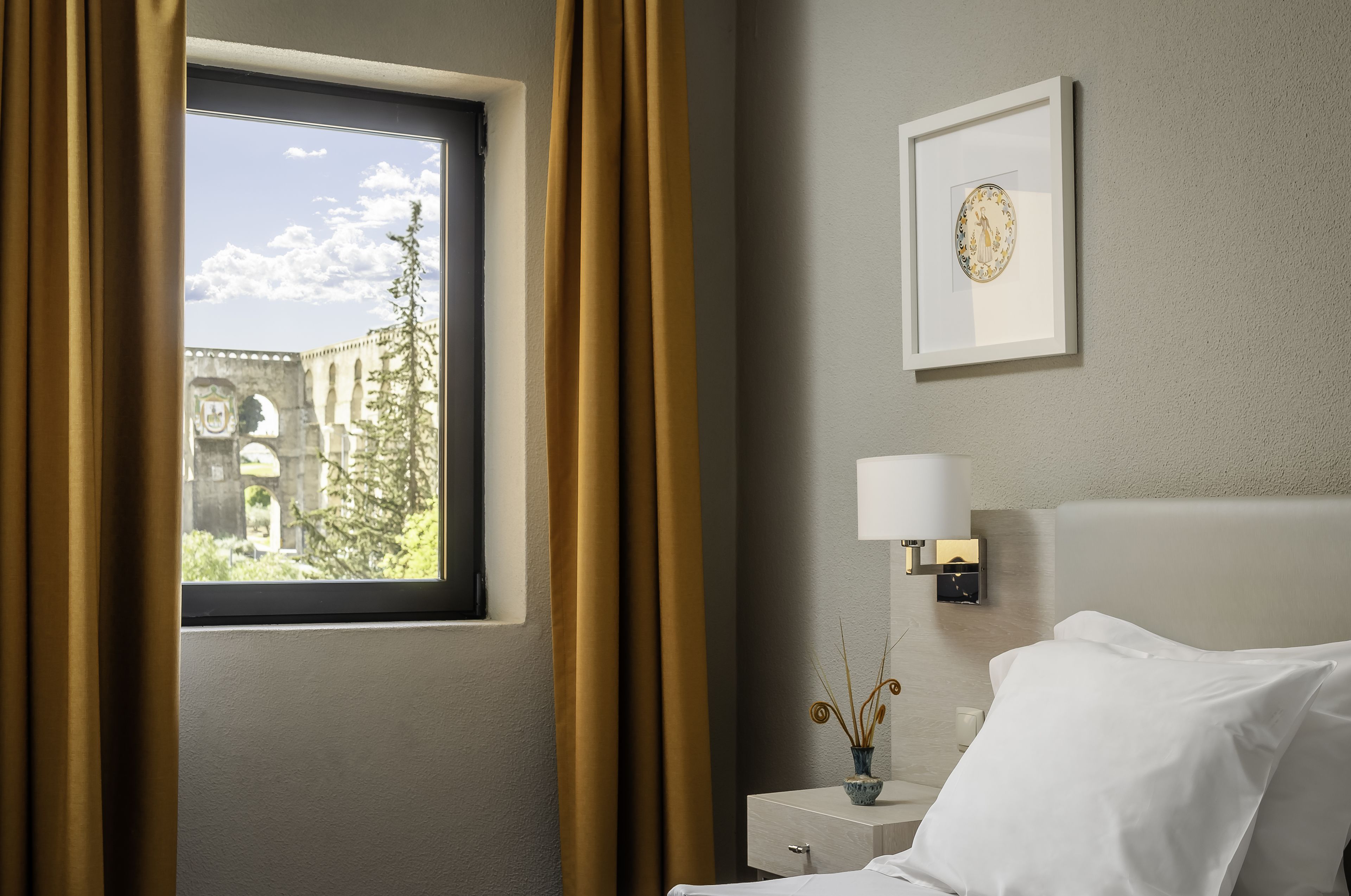 Habitación de hotel con cama, mesita de noche con lámpara, un cuadro enmarcado en la pared y una ventana con cortinas.