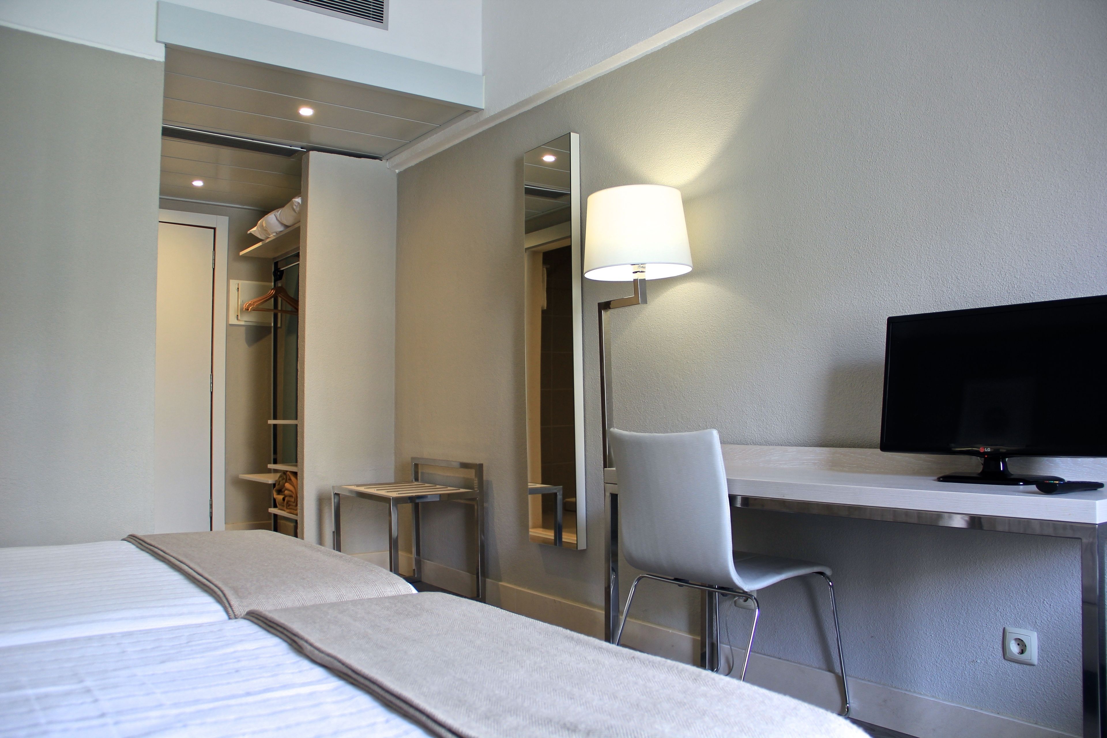 Habitación de hotel con cama, escritorio, silla, televisor y espejo.