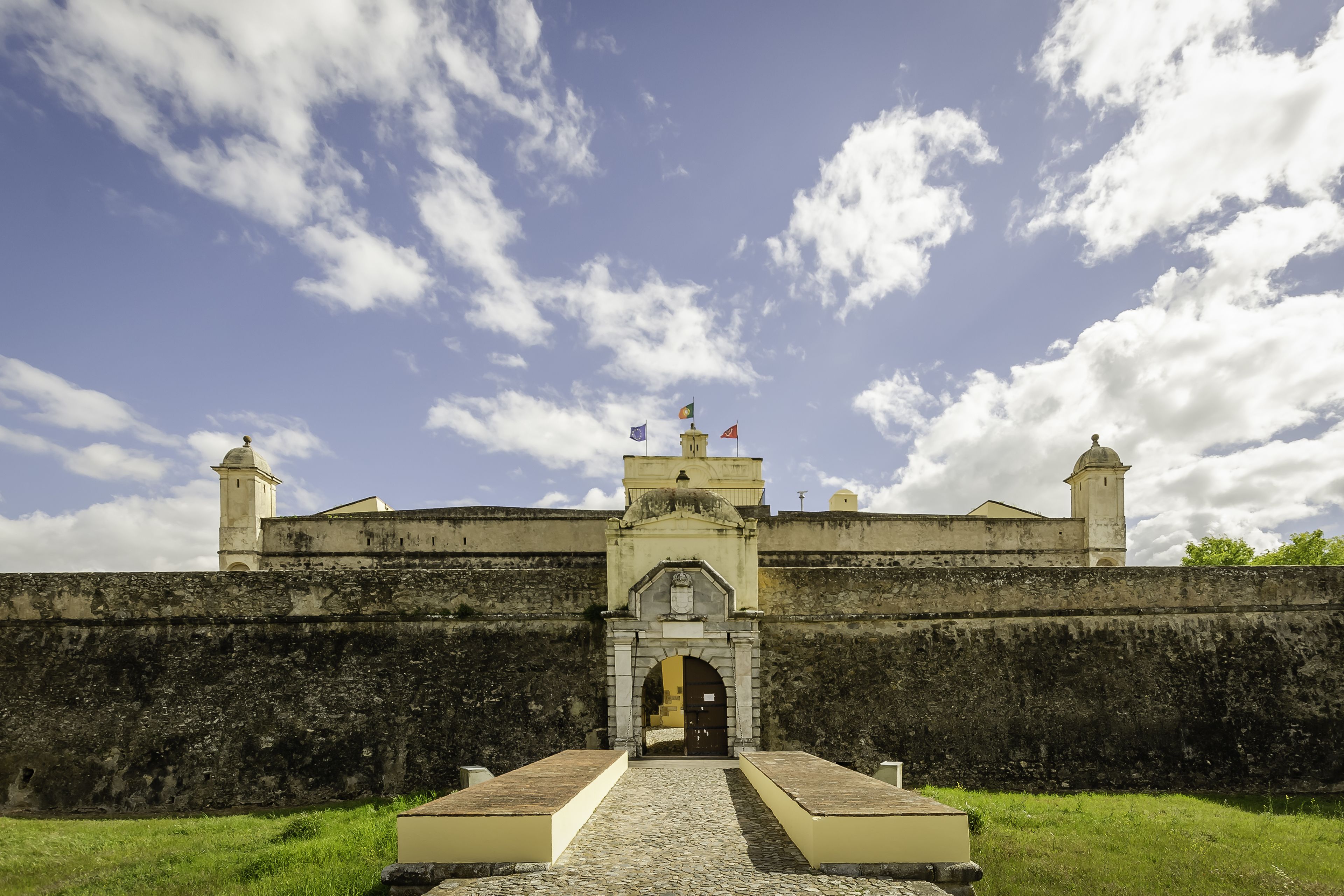 Uma fortaleza histórica com um arco central, um mastro com várias bandeiras e uma torre de vigia.