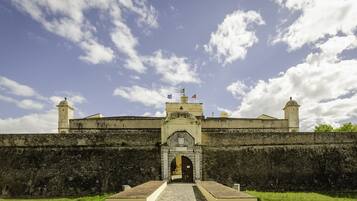 Uma fortaleza histórica com um arco central, um mastro com várias bandeiras e uma torre de vigia.