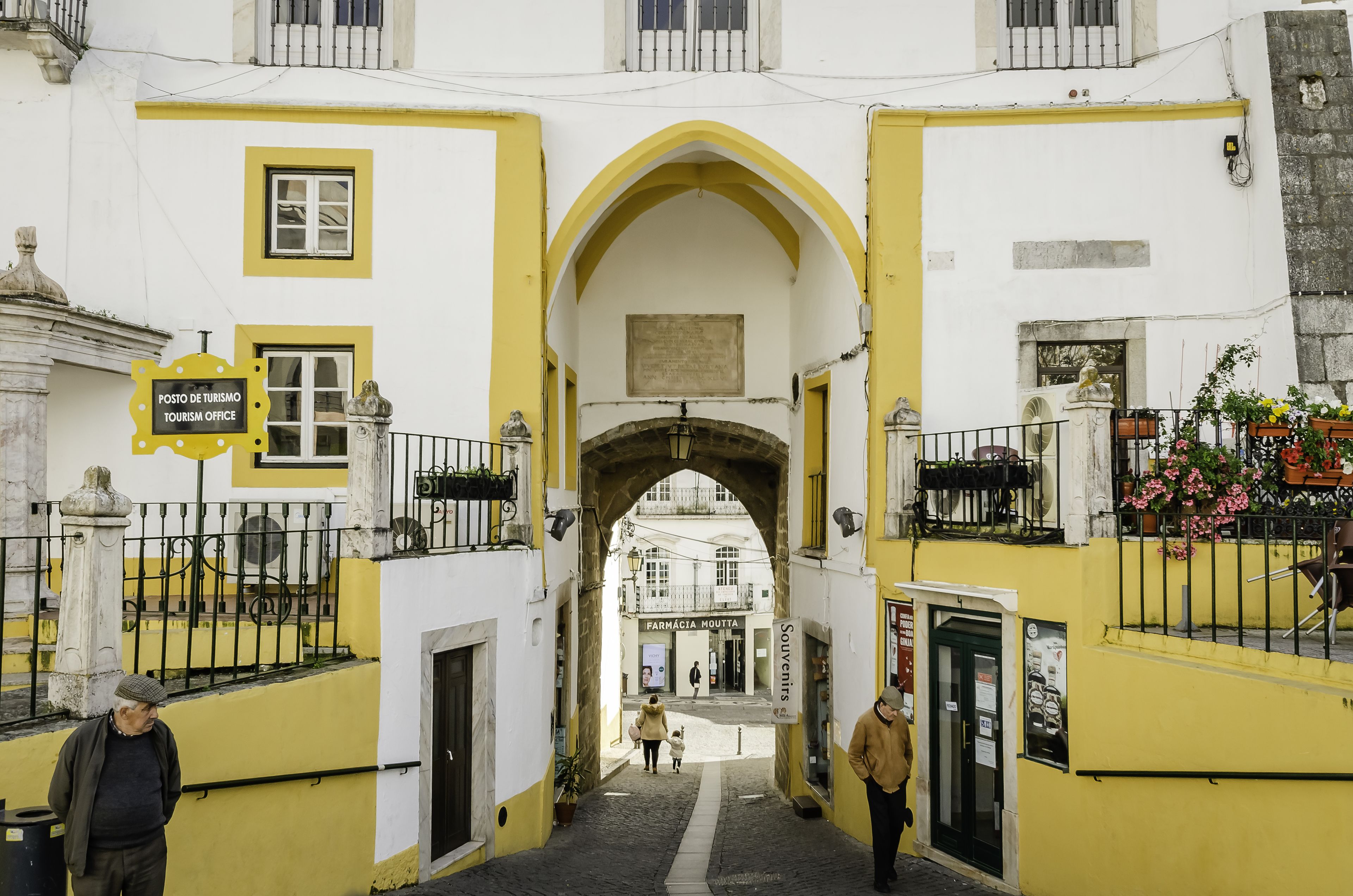 Una calle estrecha con un edificio amarillo y blanco, una farmacia y un cartel de oficina de turismo.