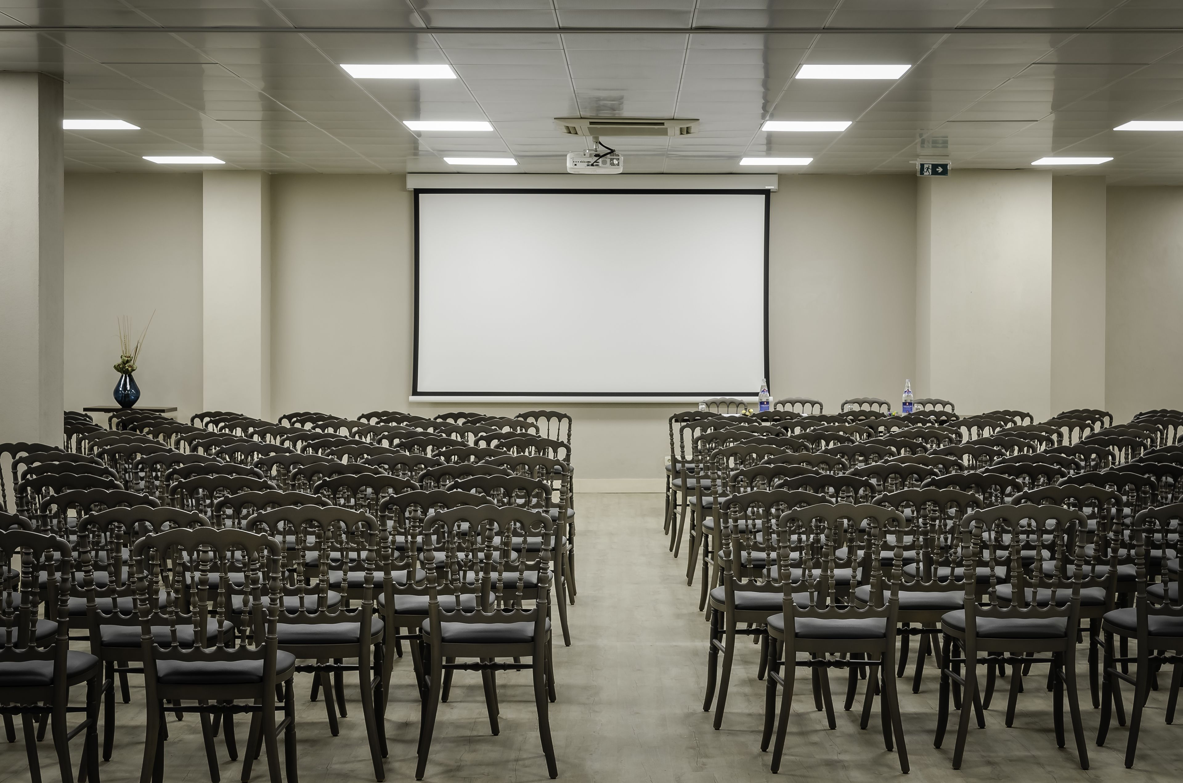 Un salón de conferencias con filas de sillas de madera orientadas hacia una pantalla de proyección grande.