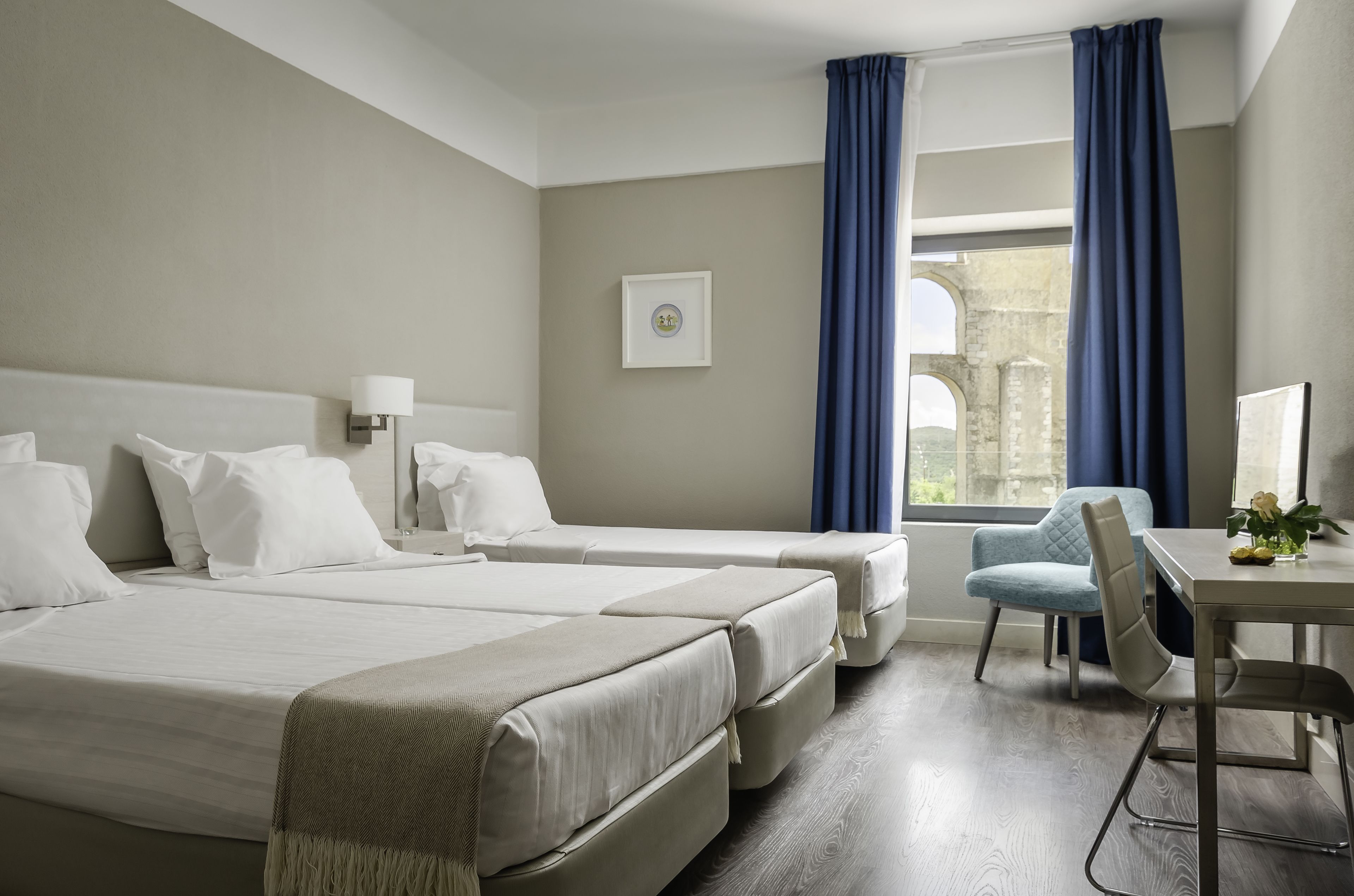 A hotel room with two beds, a desk, a chair, and a view of an old building through the window.