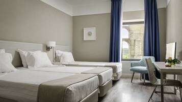 A hotel room with two beds, a desk, a chair, and a view of an old building through the window.