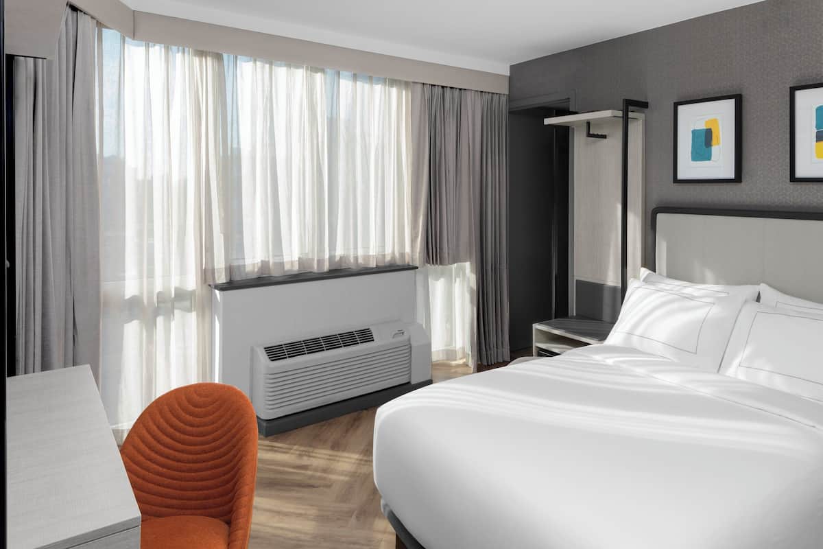 Una camera d'albergo moderna con un letto grande, una scrivania, una poltrona arancione, una finestra con tende trasparenti e un condizionatore d'aria a parete.