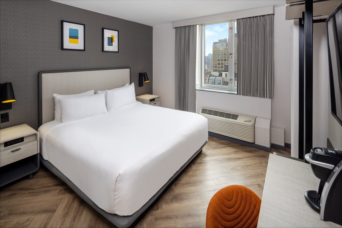 Ein Hotelzimmer mit einem großen Bett, einem Schreibtisch mit Stuhl, einem Fenster mit Vorhängen, einem Heizgerät und Bildern an der Wand.