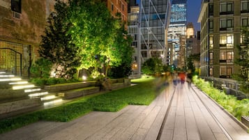 Un paseo urbano nocturno con edificios modernos, un camino bordeado de árboles y un puente.