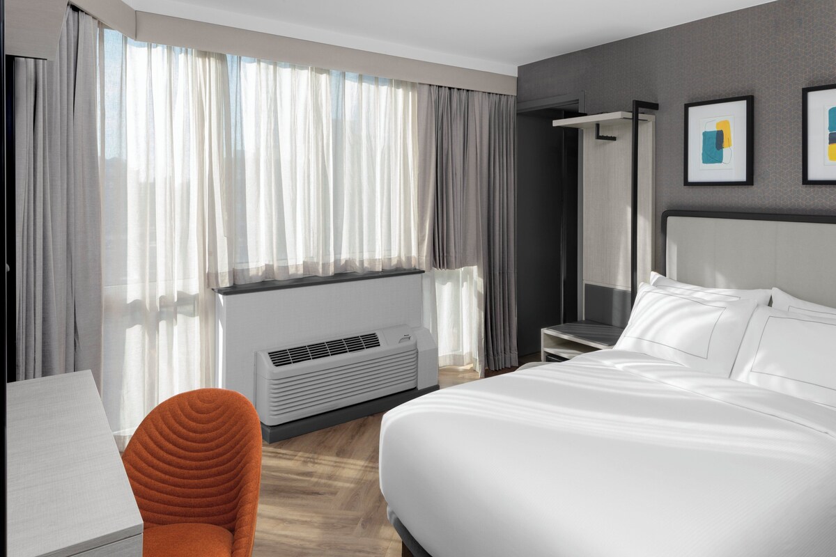 A modern hotel room with a large bed, a desk, an orange chair, a window with sheer curtains, and a wall-mounted air conditioner.