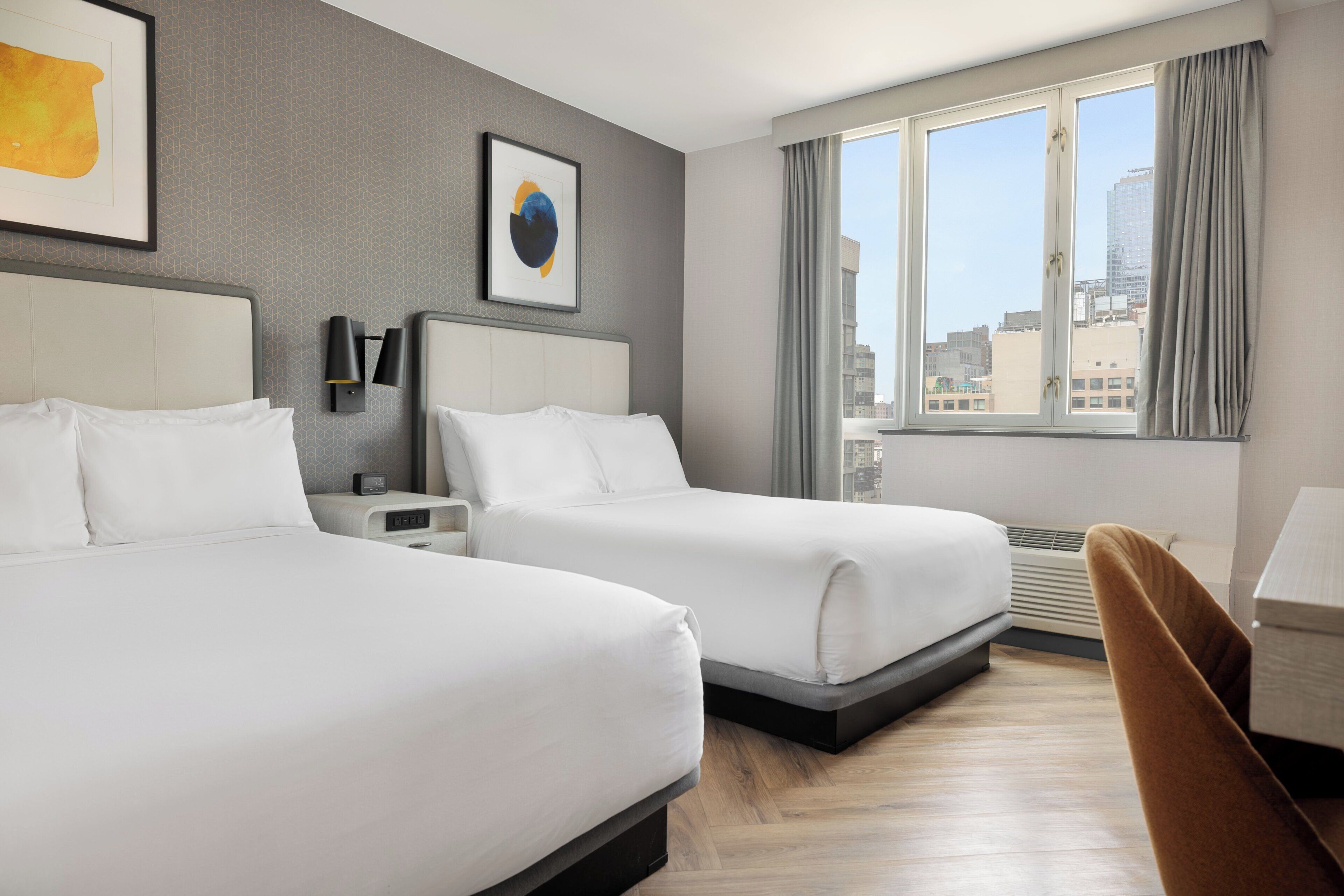Habitación de hotel con dos camas, un escritorio, una silla, un ventanal con vista a la ciudad y obras de arte en las paredes.