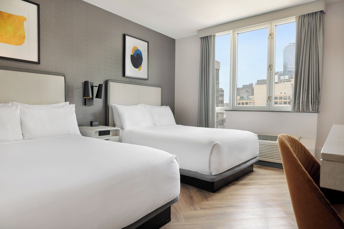 A hotel room with two beds, a desk, a chair, a window with a city view, and artwork on the walls.