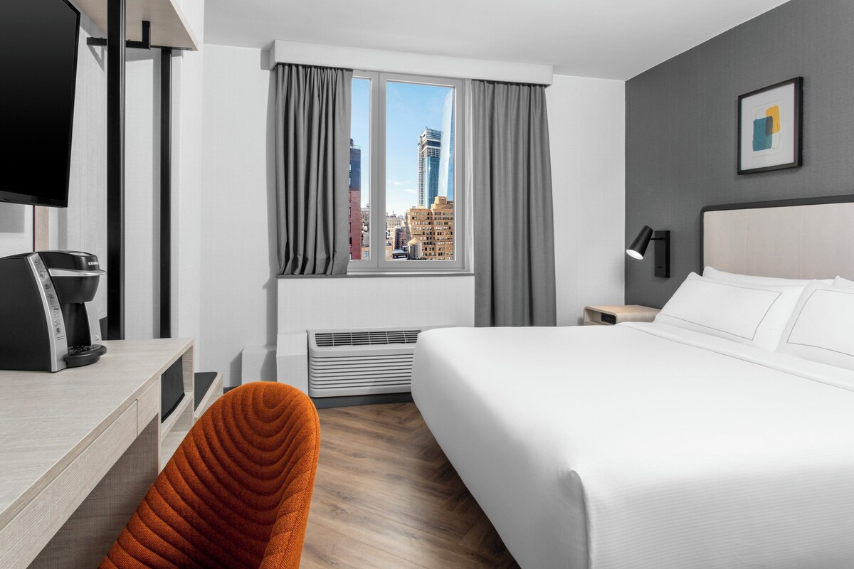 Ein Hotelzimmer mit einem Bett, einem Schreibtisch mit Kaffeemaschine, einem Sessel, einem Fenster mit Vorhängen und Blick auf die Stadt.