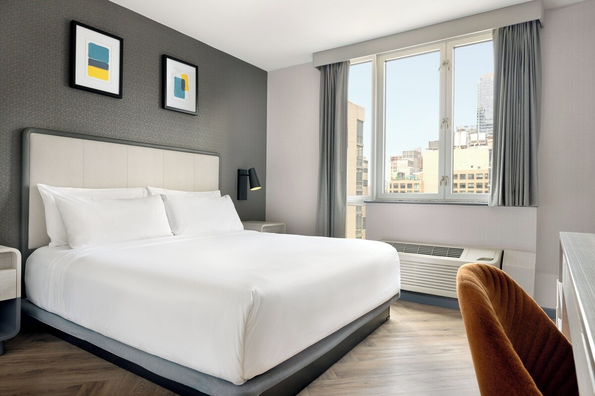 A modern hotel room with a large bed, a desk, and a window with a city view.