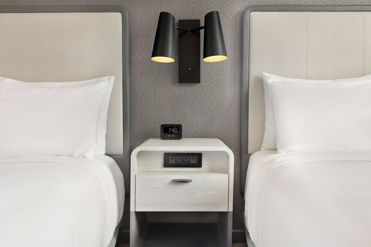 A hotel room with two beds, a nightstand with a clock, and wall-mounted lamps.
