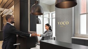 A reception desk with a modern design, a nameplate reading "VOCO AN IHG HOTEL," and a view of the interior with wooden paneling and pendant lighting.