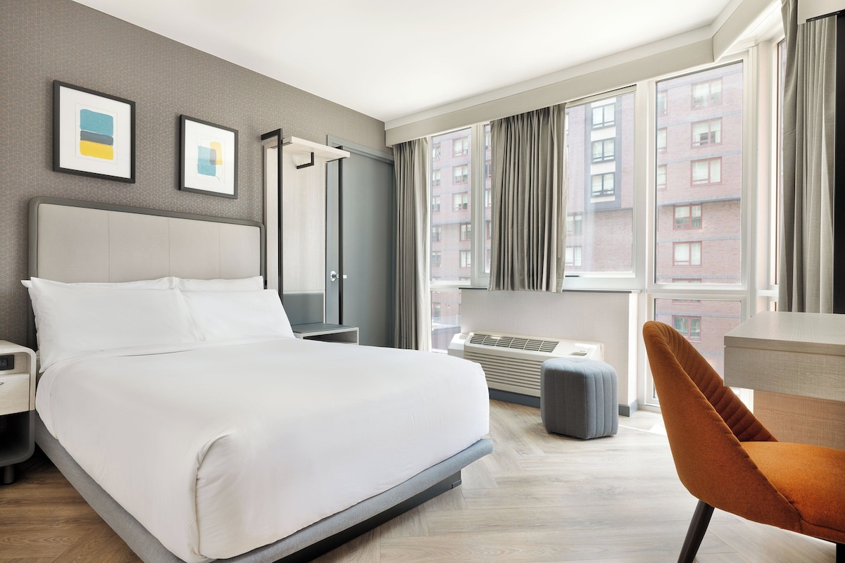 Ein modernes Hotelzimmer mit einem großen Bett, einem Schreibtisch, einem Stuhl und Blick auf eine Stadtlandschaft durch das Fenster.