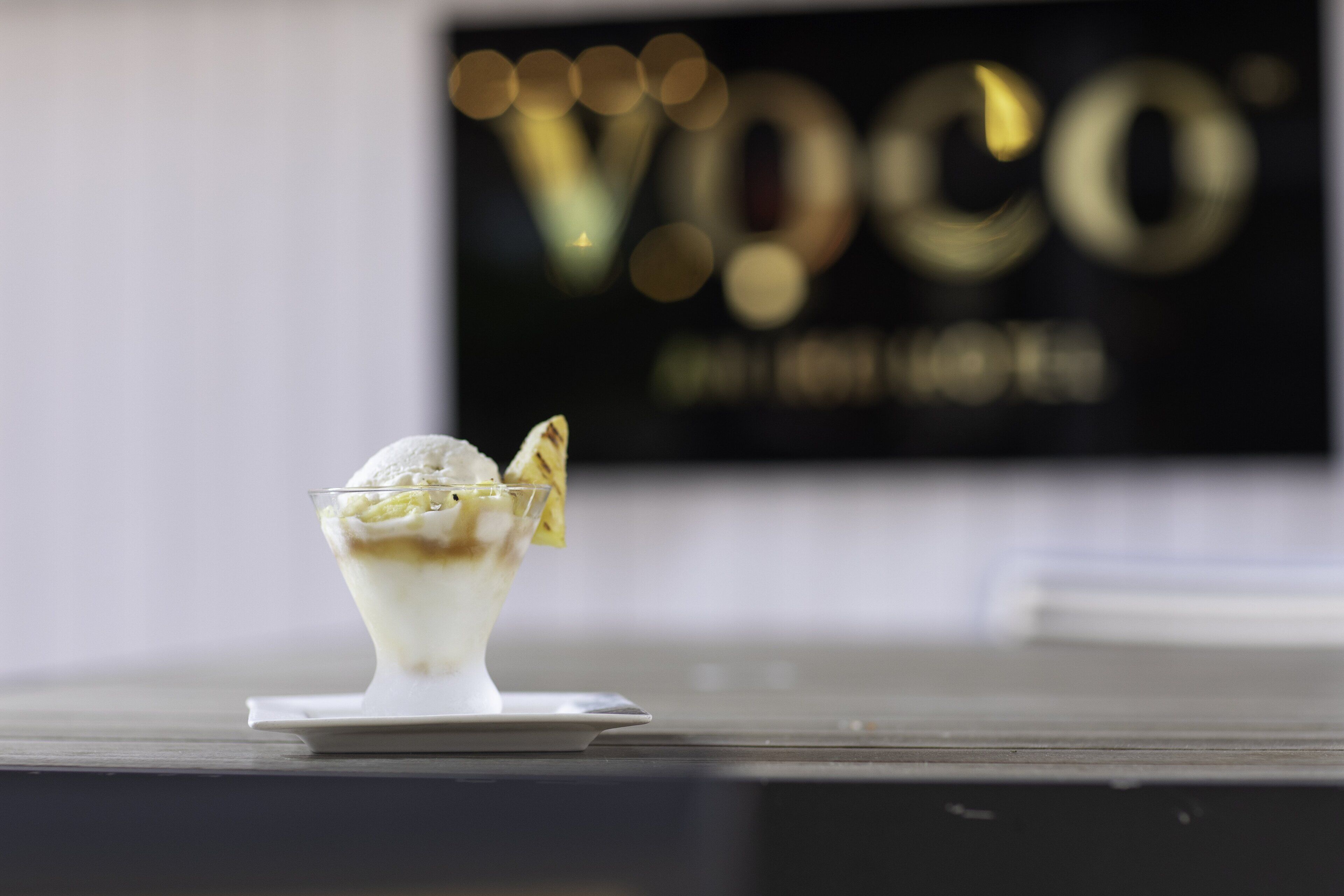 A dessert in a glass bowl on a white plate, with a blurred background featuring a sign with the letters "VPC".