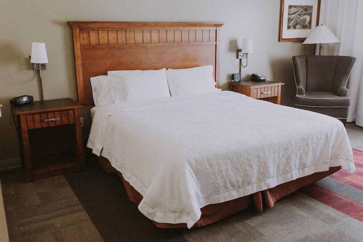 Une chambre d’hôtel avec un grand lit, une tête de lit en bois, des tables de chevet, une chaise et une lampe.