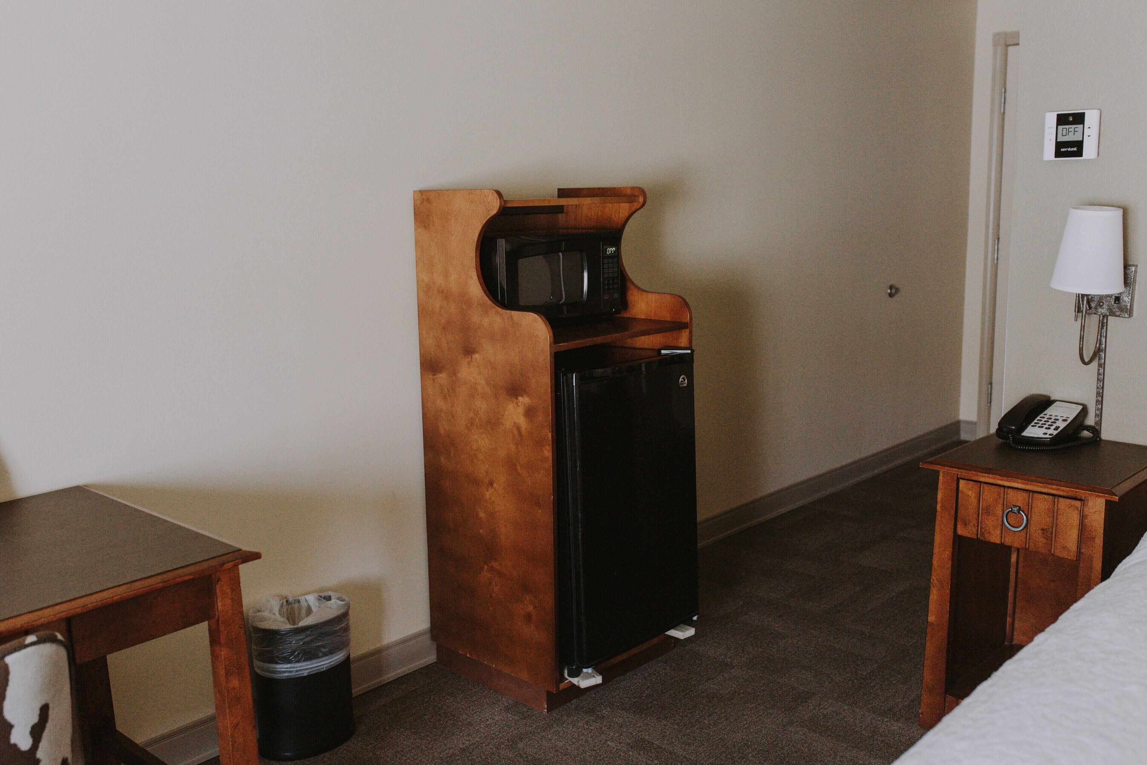 Habitación de hotel compacta con escritorio de madera, un refrigerador pequeño, un cesto de basura, una mesita de noche con lámpara y teléfono en la pared.
