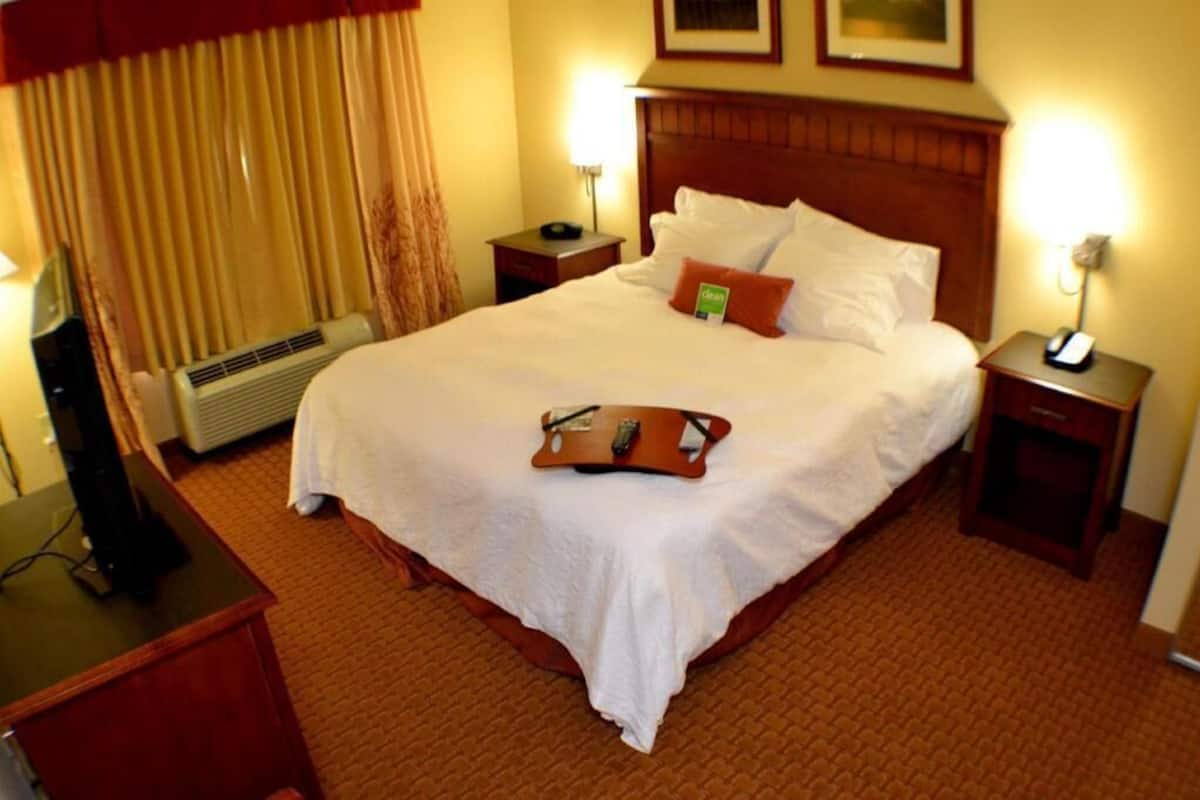Une chambre d’hôtel avec un grand lit, deux tables de chevet, un bureau avec une chaise, un radiateur et une fenêtre avec des rideaux.