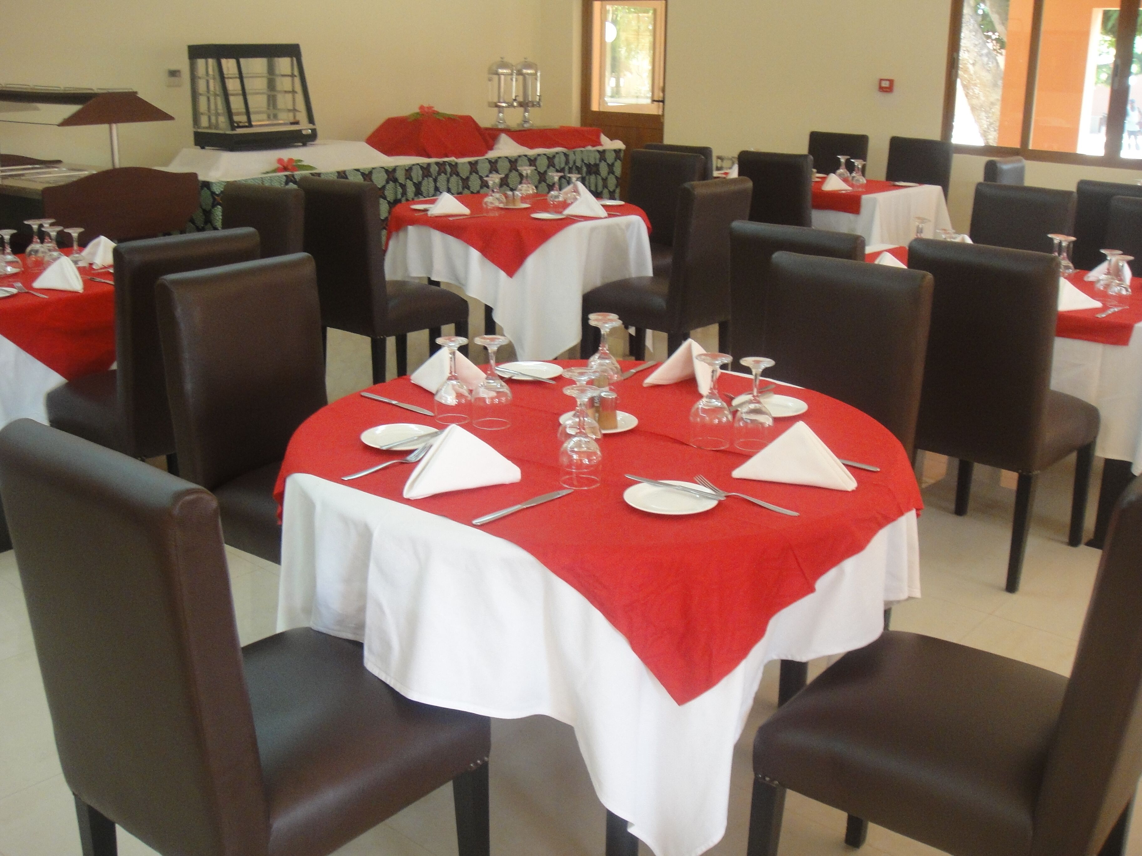 Un comedor con mesas redondas cubiertas con manteles rojos, cada una con servilletas blancas, cubiertos y copas de vino.