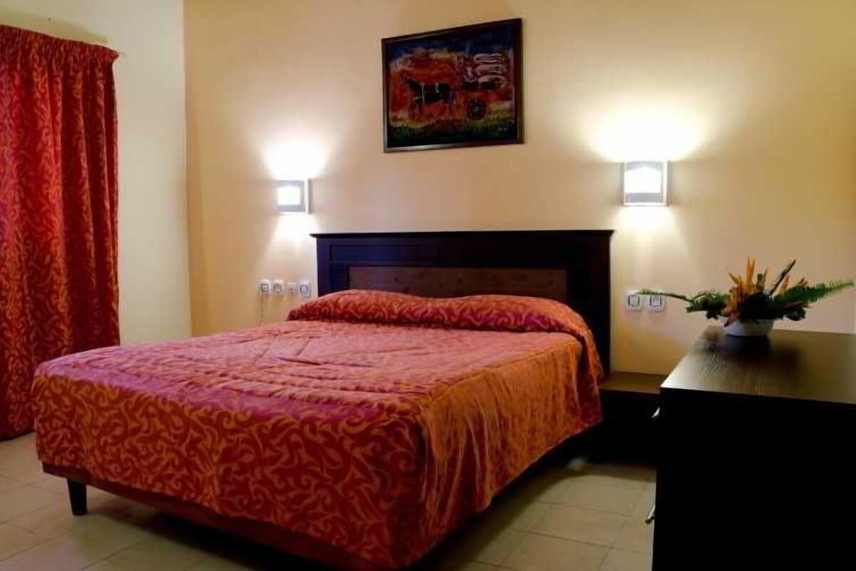 A bedroom with a bed, two wall-mounted lights, a painting, and a plant on a table.