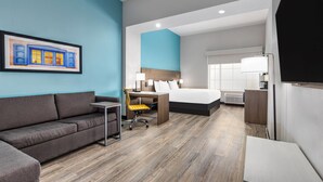 A modern hotel room with a gray sofa, a desk with a chair, a bed, and a large window. - La Quinta Inn & Suites by Wyndham Hobbs (Hobbs)