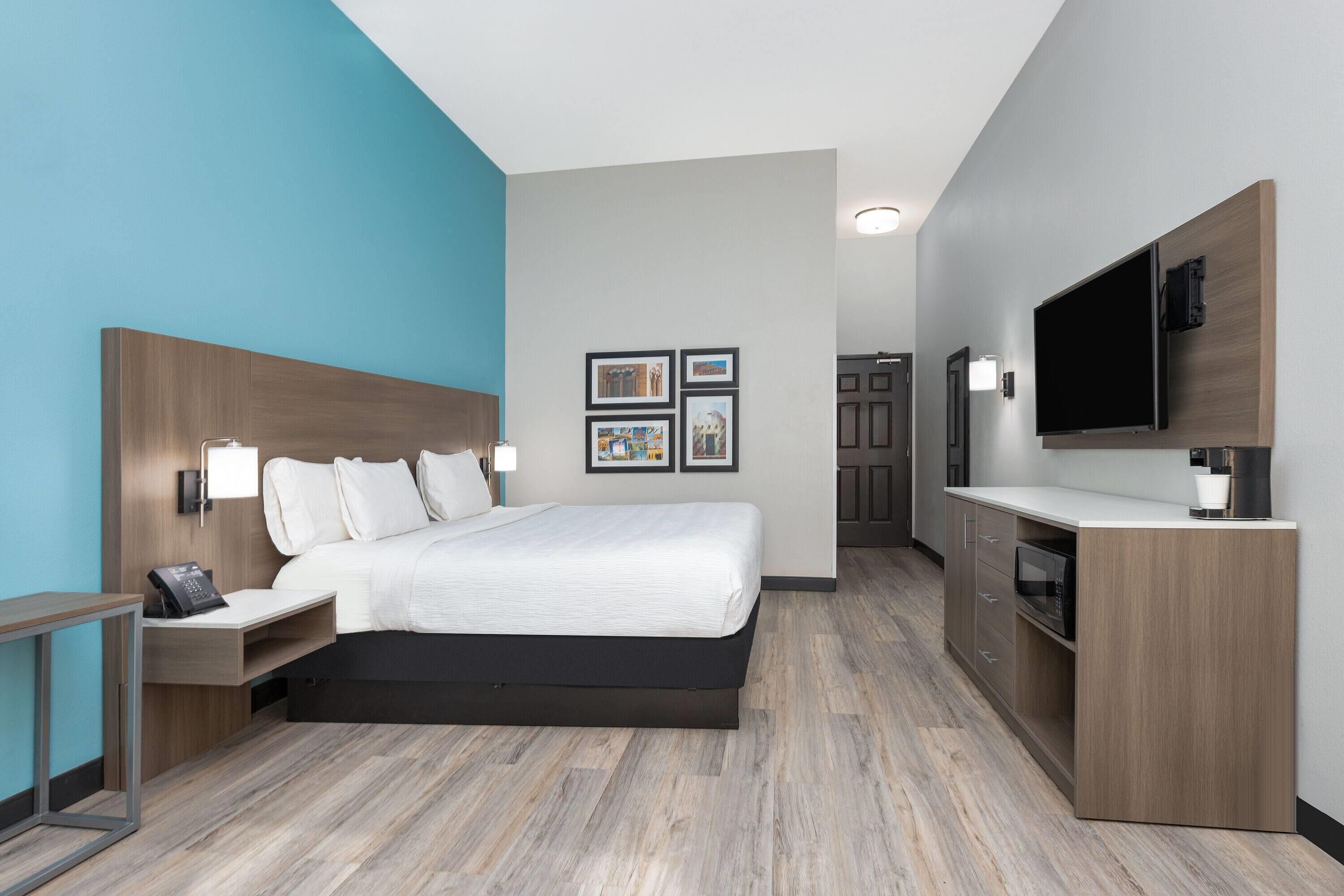A modern hotel room with a large bed, a desk with a phone, a flat-screen TV, and a kitchenette.