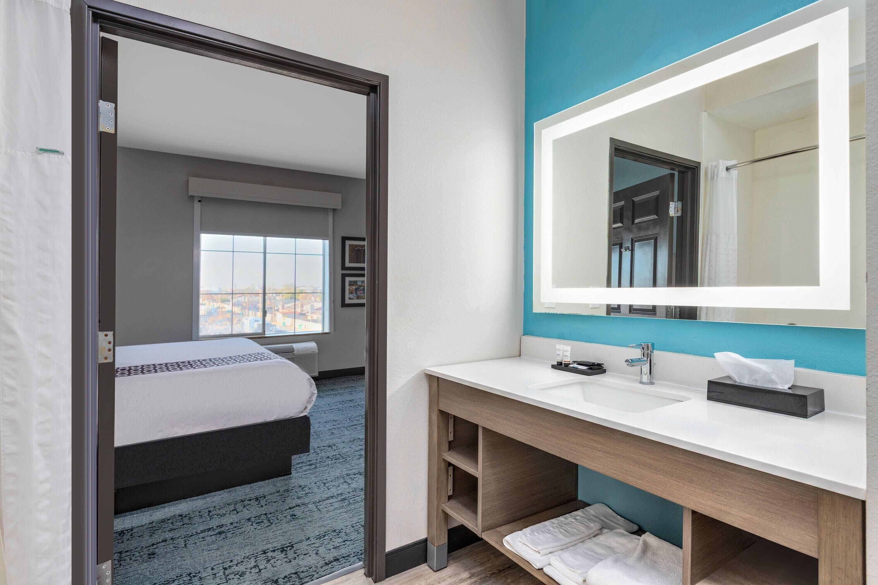 Habitación de hotel moderna con una cama grande, un baño con lavamanos y espejo blancos, y vistas de la ciudad a través de la ventana.
