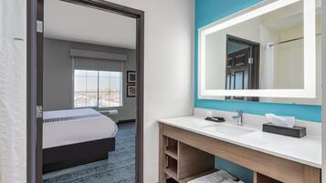 Habitación de hotel moderna con una cama grande, un baño con lavamanos y espejo blancos, y vistas de la ciudad a través de la ventana.