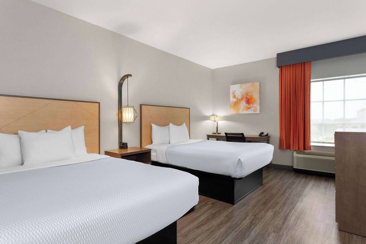 Een hotelkamer met twee bedden, een bureau, een stoel, een lamp, een schilderij en een raam met gordijnen.
