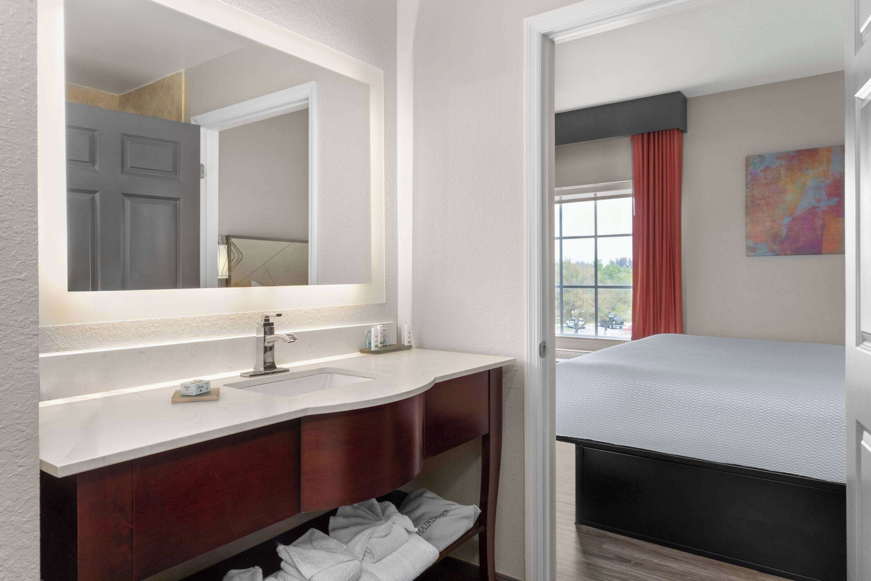 A hotel room with a large mirror, a sink, a vanity, a bed, and a window with curtains.
