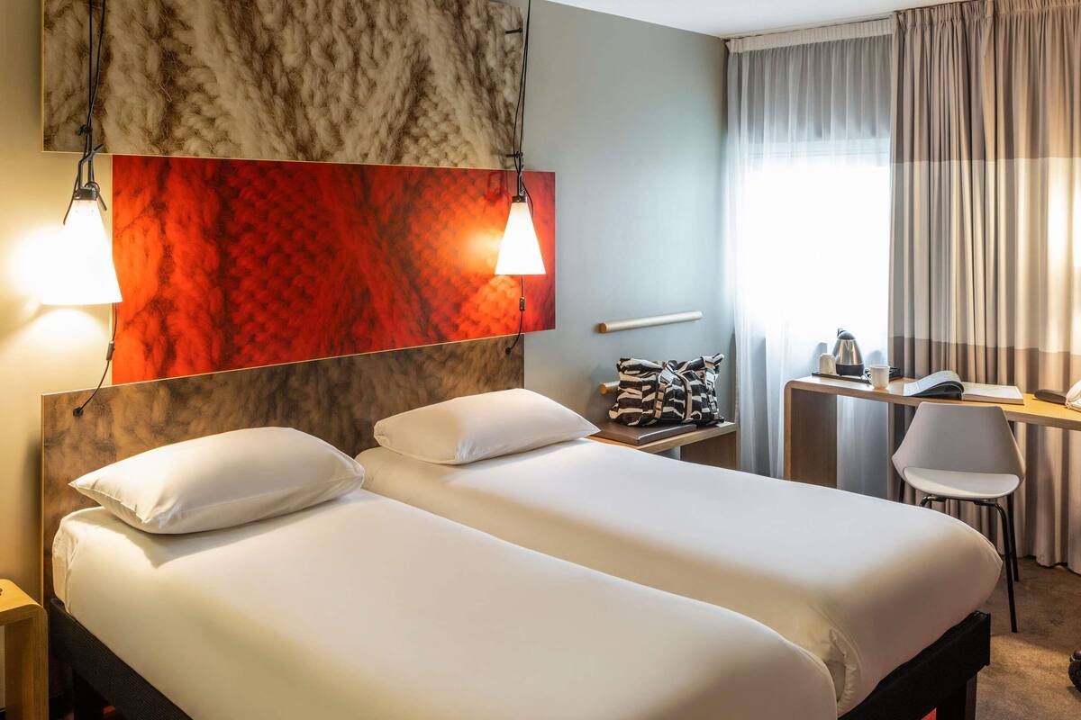 Habitación de hotel con dos camas, un escritorio, una silla, una lámpara y un ventanal con cortinas.
