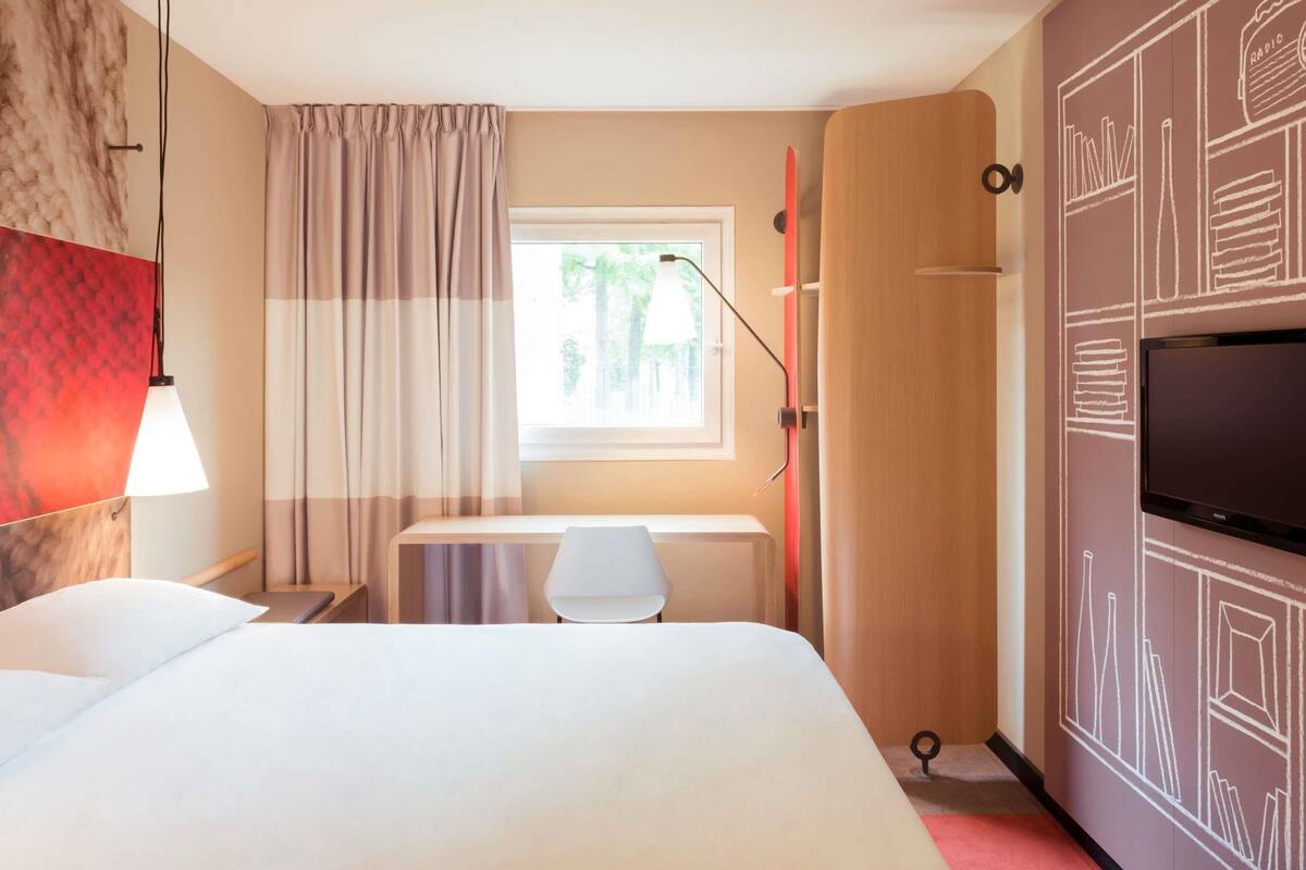 Una habitación de hotel con cama, escritorio, silla, televisión, ventana con cortinas y pared con diseño geométrico.
