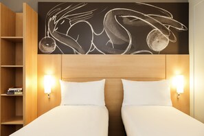 Habitación de hotel con cabecera de madera, dos almohadas blancas, una lámpara de pared y una estantería con libros.