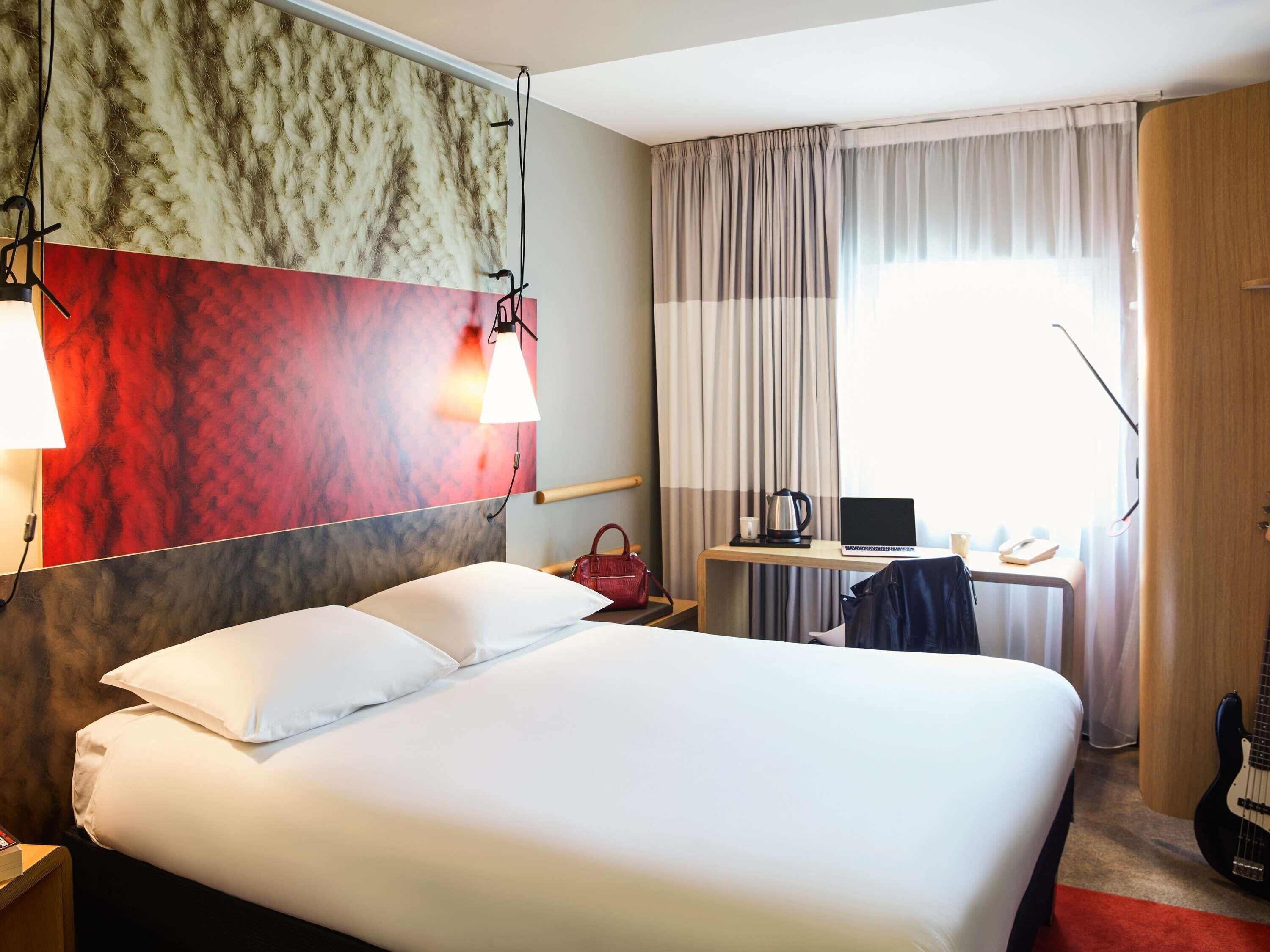 A hotel room with a large bed, a desk with a laptop, a chair, a guitar, and a window with curtains.