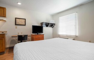 Un dormitorio con cama, escritorio con computadora, televisión y ventana con persianas.