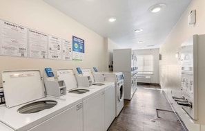Une laverie équipée de plusieurs machines à laver et de plusieurs séchoirs, avec des affiches explicatives accrochées au mur et un aménagement propre et moderne.