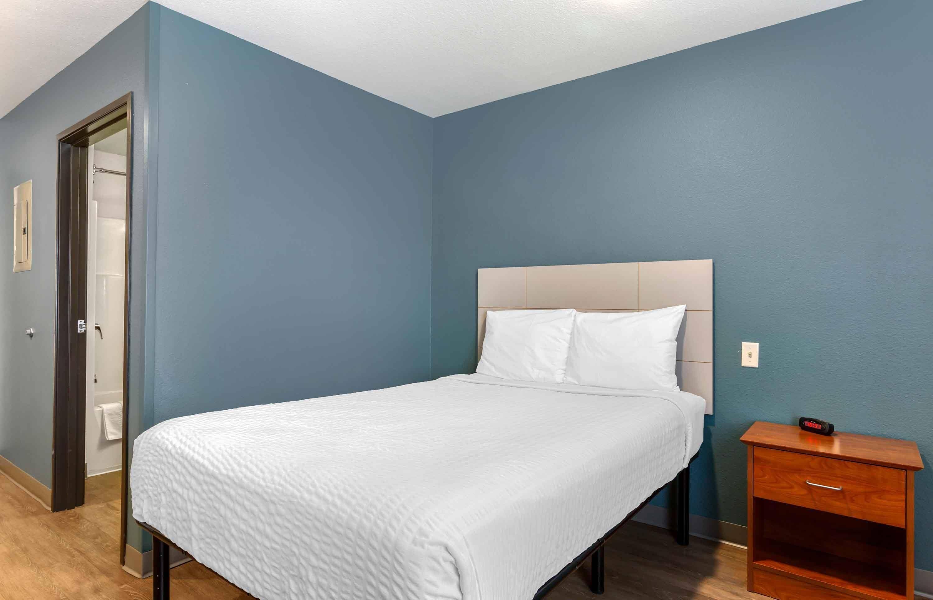 A neatly made bed in a room with blue walls, a wooden nightstand, and a door leading to a bathroom.