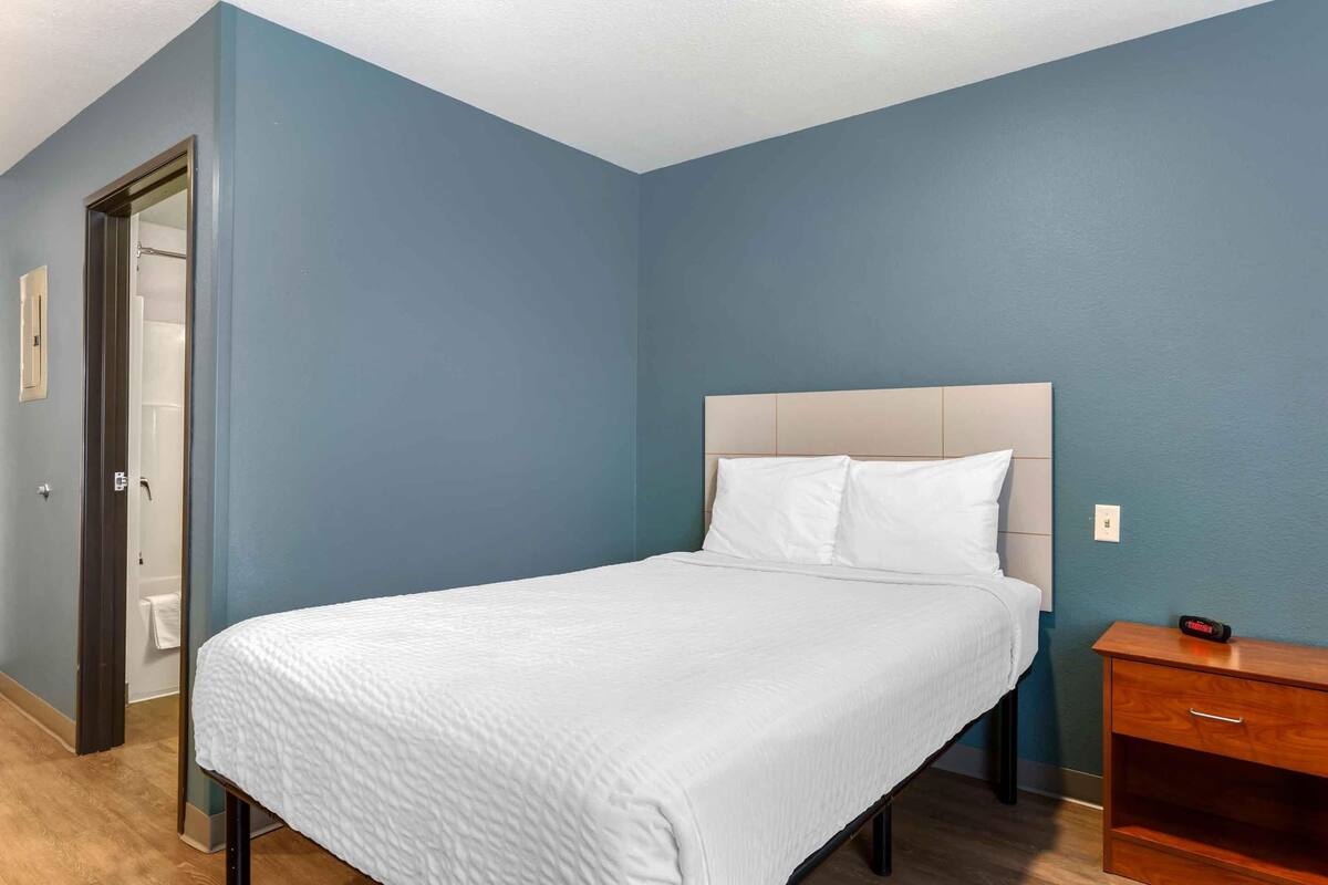 Un lit bien fait dans une pièce aux murs bleus, avec une table de chevet en bois et une porte donnant accès à une salle de bain.