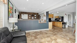 A modern living room with a black leather couch, a kitchen area with wooden cabinets, and a tiled floor.