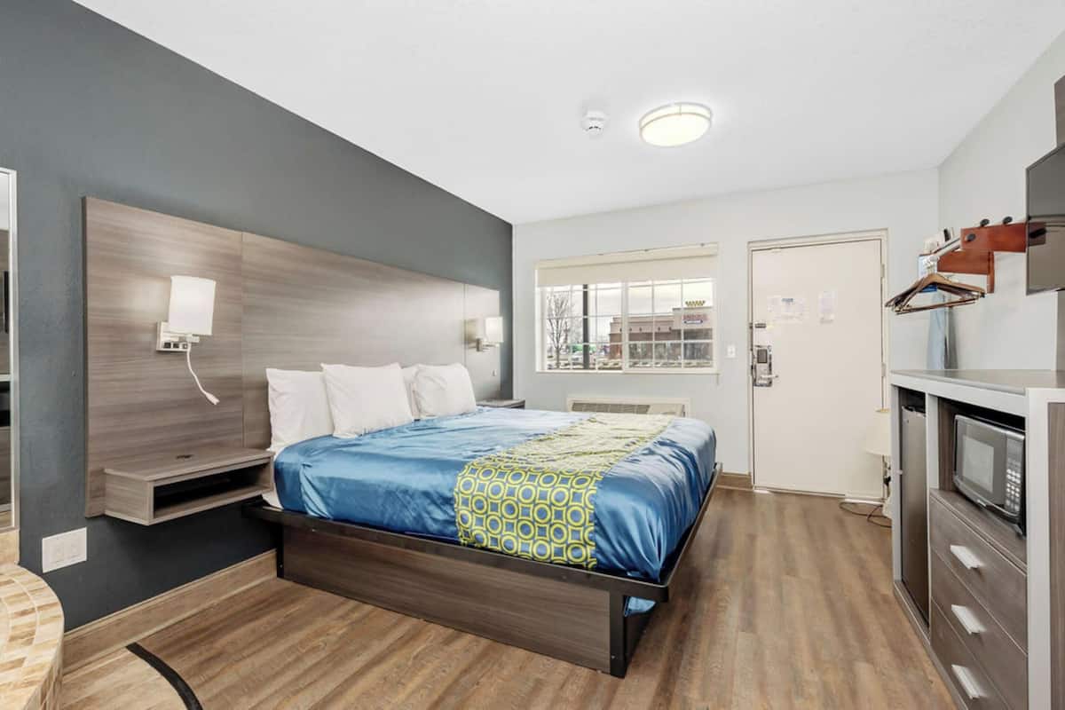 A modern hotel room with a wooden bed, a built-in TV, a kitchenette, and a window with a city view.