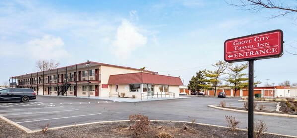 A travel inn with a red entrance sign, a parking lot, and a building with multiple floors and a red roof. - Grove City Travel Inn Columbus South (Grove City)